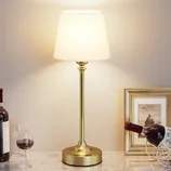 A table lamp with a gold base and a white shade is placed on a marble surface, surrounded by wine bottles, a wine glass, and a framed picture.