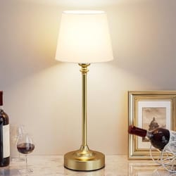 A table lamp with a gold base and a white shade is placed on a marble surface, surrounded by wine bottles, a wine glass, and a framed picture.