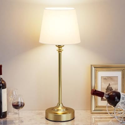 A table lamp with a gold base and a white shade is placed on a marble surface, surrounded by wine bottles, a wine glass, and a framed picture.