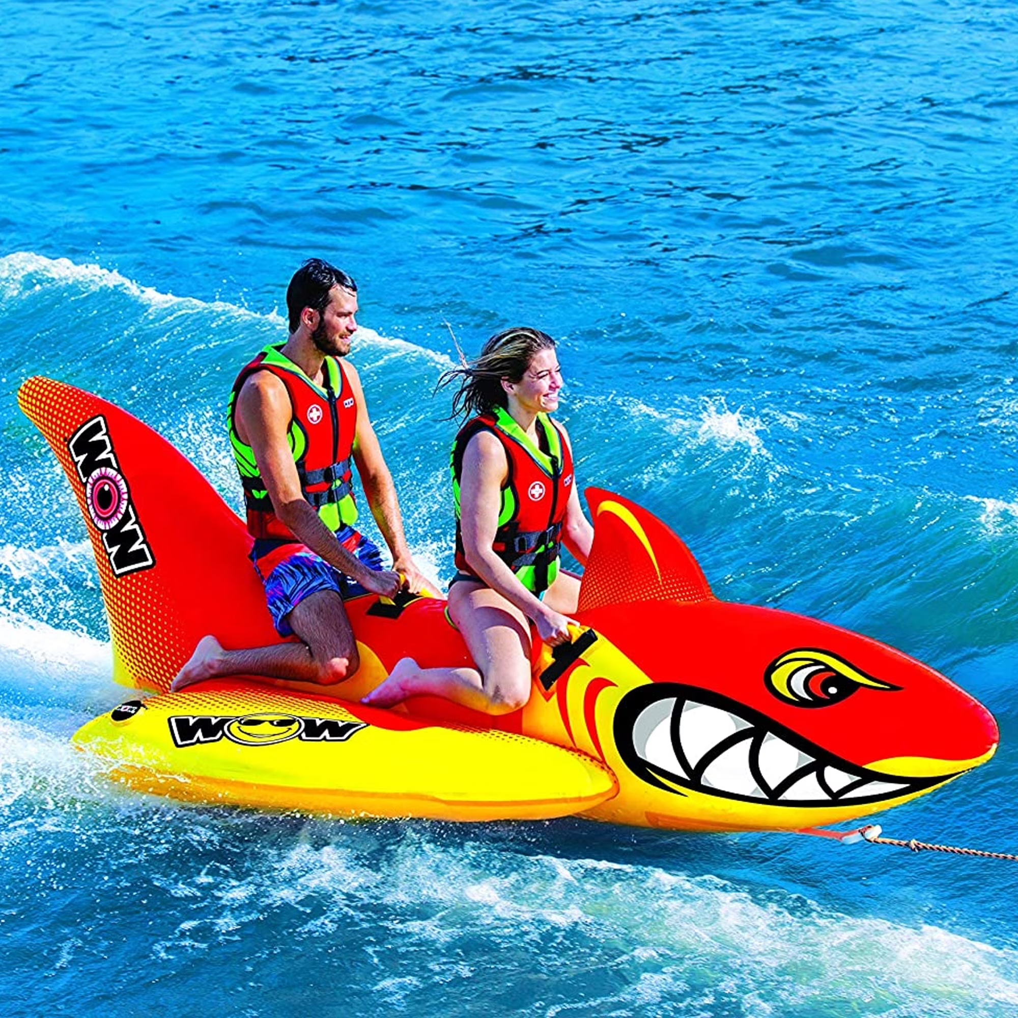 An inflatable towable water sports raft shaped like a shark, designed to hold two people. The riders wear life vests and are positioned on the raft, which is being pulled across the water.