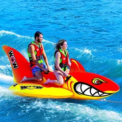 An inflatable towable water sports raft shaped like a shark, designed to hold two people. The riders wear life vests and are positioned on the raft, which is being pulled across the water.