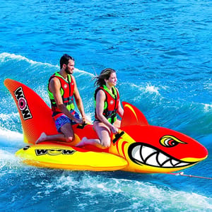 An inflatable towable water sports raft shaped like a shark, designed to hold two people. The riders wear life vests and are positioned on the raft, which is being pulled across the water.