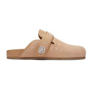 Coach Outlet Suede Clogs with Rivets are tan slip-on shoes featuring a suede upper with small rivet accents and a metallic logo clasp on the adjustable strap. They have a flat sole for comfort.