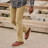 A person wearing khaki pants and red-brown leather boots with a crepe sole walks on a path, with a plaid shirt visible above. The scene includes a wooden railing and rocky background.