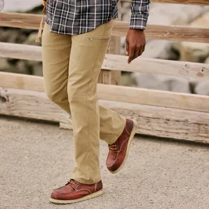 A person wearing khaki pants and red-brown leather boots with a crepe sole walks on a path, with a plaid shirt visible above. The scene includes a wooden railing and rocky background.