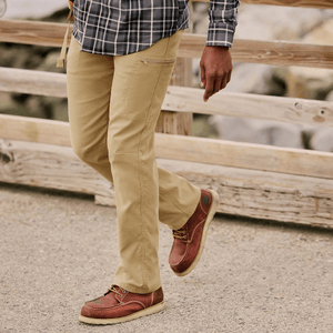 A person wearing khaki pants and red-brown leather boots with a crepe sole walks on a path, with a plaid shirt visible above. The scene includes a wooden railing and rocky background.
