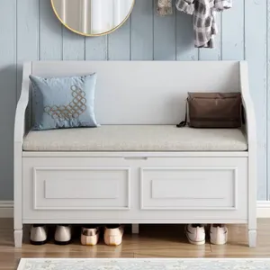A white storage bench with a cushioned seat holds a decorative pillow and a brown bag, with shoes neatly placed underneath. The light blue paneled wall features a round mirror and hanging clothes.