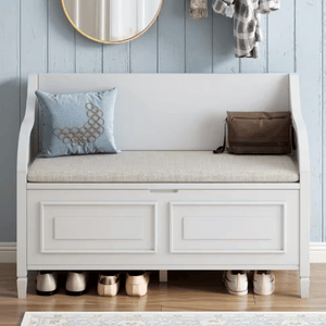 A white storage bench with a cushioned seat holds a decorative pillow and a brown bag, with shoes neatly placed underneath. The light blue paneled wall features a round mirror and hanging clothes.