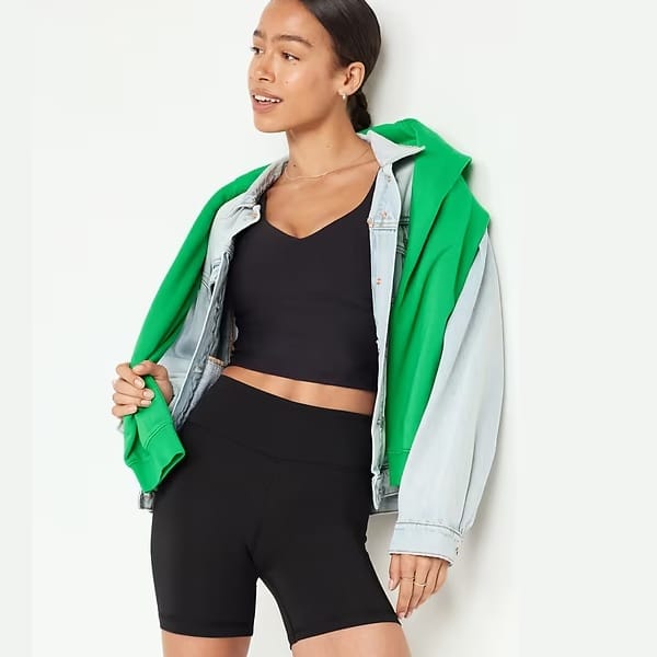 A woman is wearing a black crop top, black shorts, a green hooded jacket, and a light blue denim jacket draped over her shoulders.