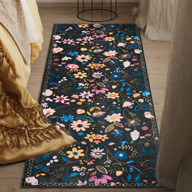 Floral-patterned rug featuring colorful flowers on a dark background, placed beside a bed with a yellow fringed blanket.