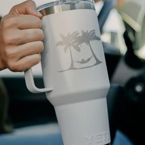 A person is holding a white YETI travel mug featuring a palm tree and hammock design.