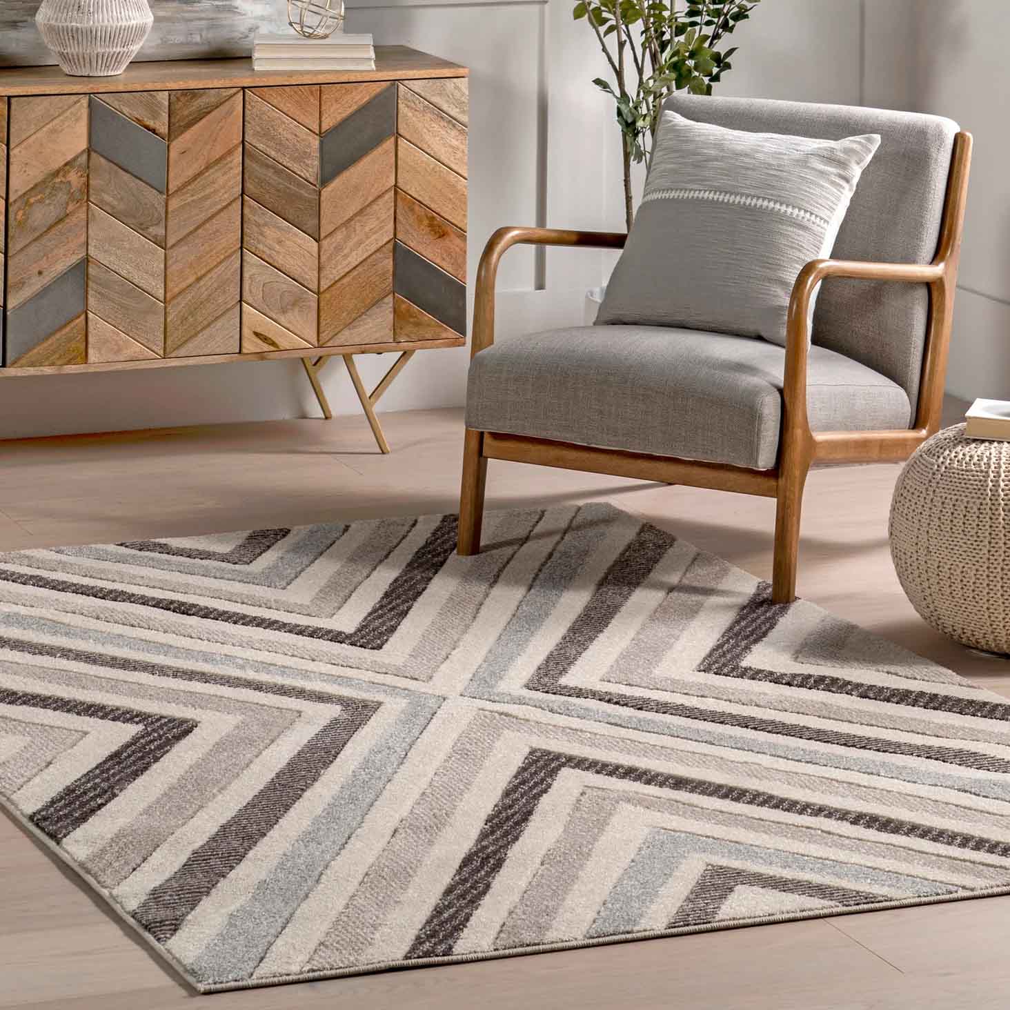 A 5' x 8' NuLoom Chevron Area Rug featuring a geometric pattern in beige, gray, and brown tones, paired with a light gray armchair and a woven pouf, enhancing a modern, minimalist living space.