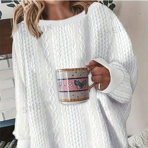 A person is wearing a white cable knit sweater and holding a mug with a plaid pattern and butterfly motif.