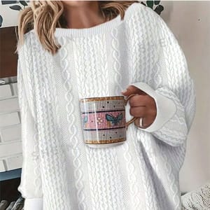 A person is wearing a white cable knit sweater and holding a mug with a plaid pattern and butterfly motif.