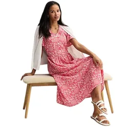 A woman in a pink floral dress sits on a bench, draped with a white cardigan. She wears white strappy sandals.