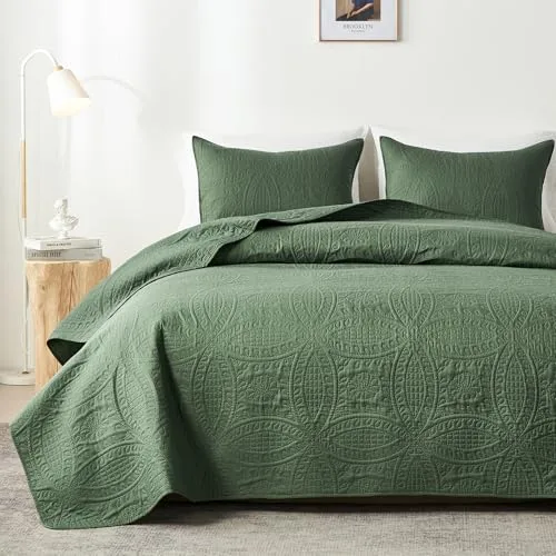 An olive green quilt with an intricate geometric pattern is neatly arranged on a bed with matching pillow shams, alongside a modern lamp on a wooden side table.