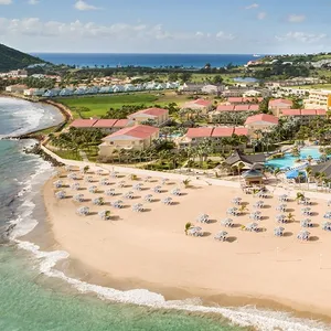 Aerial view of a beachfront resort with numerous umbrellas and loungers on the sandy shore, adjacent to multiple buildings with red roofs, lush greenery, and a large swimming pool.