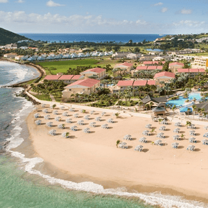 Aerial view of a beachfront resort with numerous umbrellas and loungers on the sandy shore, adjacent to multiple buildings with red roofs, lush greenery, and a large swimming pool.