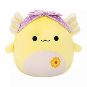 A yellow Squishmallow axolotl with a cream belly and sunflower detail, features fluffy gills, a simple smiley face, and wears a purple polka-dotted bow on its head.
