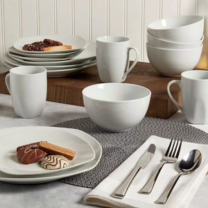 A set of white ceramic dinnerware including plates, bowls, and mugs, accompanied by a silverware set on a napkin. Cookies are placed on a plate, all arranged on a wooden surface with a textured placemat.