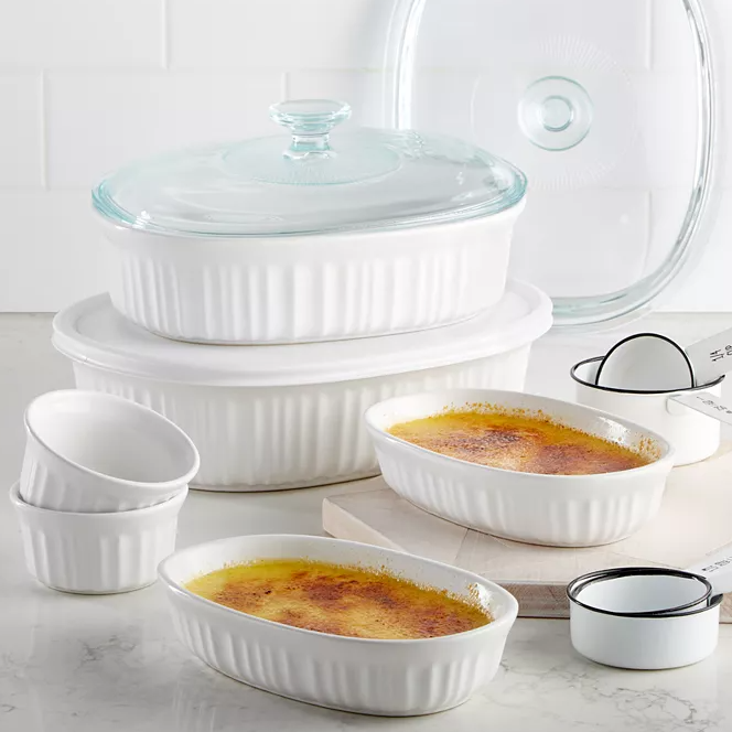 A collection of white, ribbed ceramic bakeware, including round and oval dishes with glass lids, ramekins, and small bowls, displayed on a kitchen countertop.