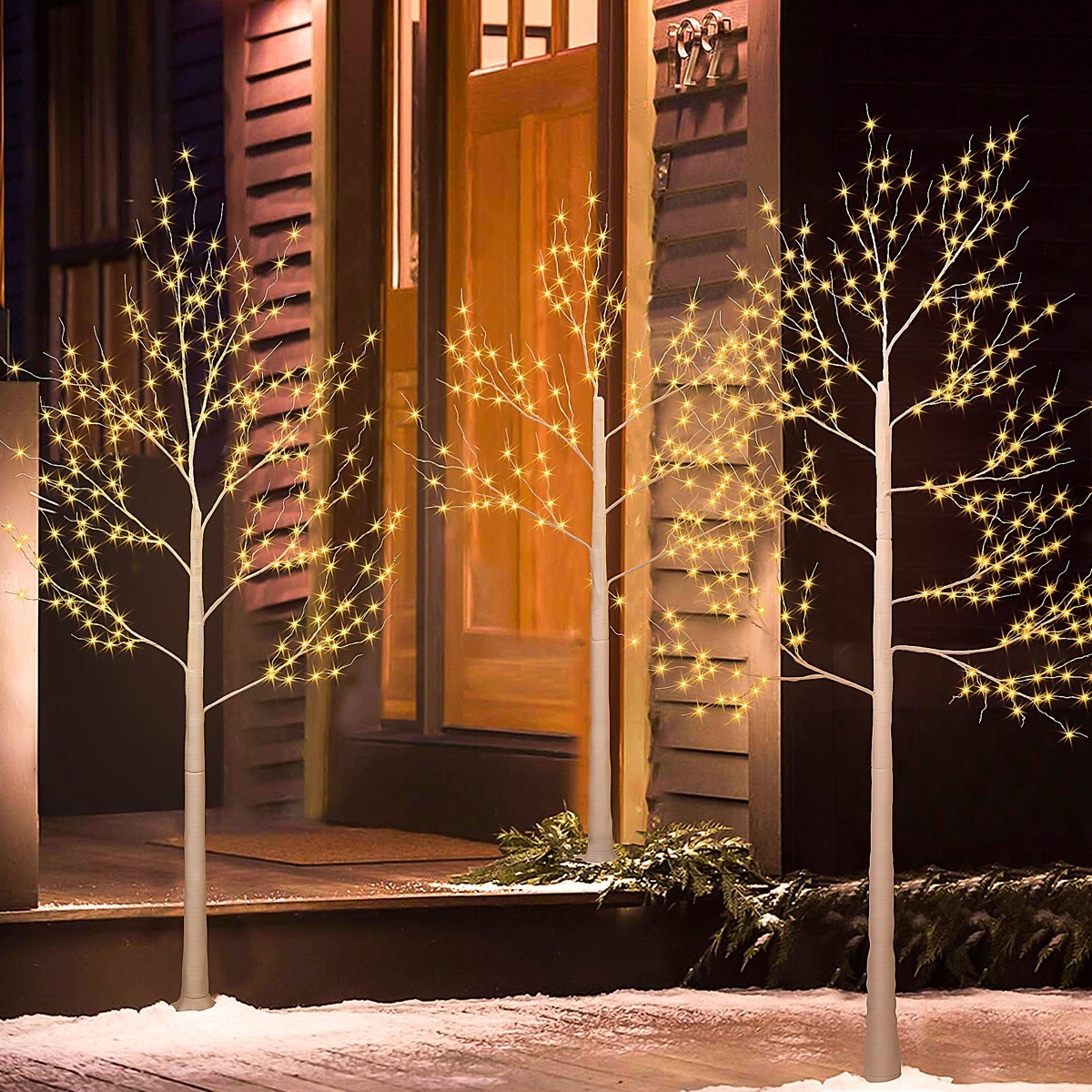 LED lighted artificial trees with warm white lights, displayed on a snowy outdoor porch, creating a festive ambiance near a wooden door.