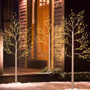 LED lighted artificial trees with warm white lights, displayed on a snowy outdoor porch, creating a festive ambiance near a wooden door.