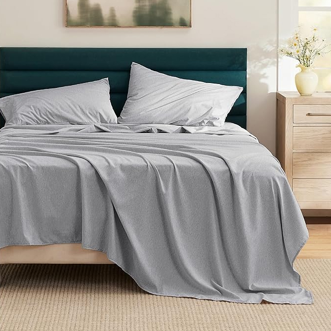 A neatly made bed features light gray deep-pocket microfiber sheets with matching pillowcases. The bed has a dark green upholstered headboard, a beige bed frame, and sits atop a light brown woven rug.