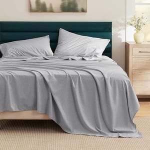 A neatly made bed features light gray deep-pocket microfiber sheets with matching pillowcases. The bed has a dark green upholstered headboard, a beige bed frame, and sits atop a light brown woven rug.