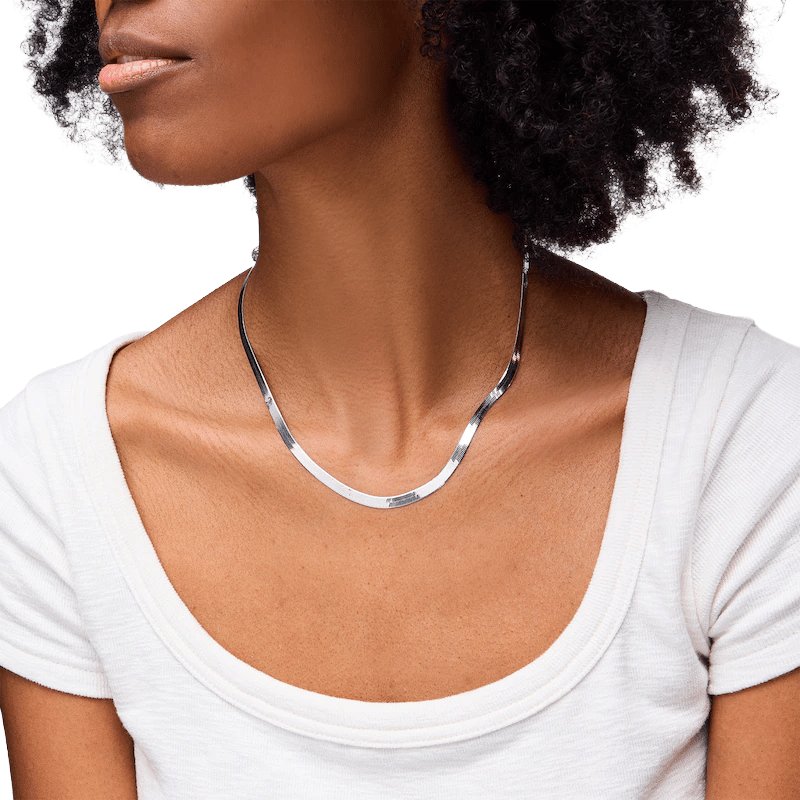 A woman wearing a minimalist, shiny silver necklace with a sleek, curved design, paired with a white scoop-neck top.