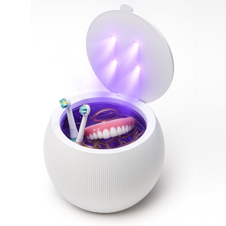 A UV sterilizer for dental items, containing a pair of dentures, electric toothbrush heads, and a golden ring, is opened with four UV lights visible inside.