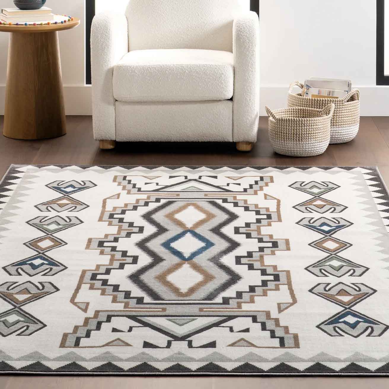 NuLoom Geometric Machine Washable Rug features a tribal-inspired pattern in neutral tones of white, brown, and blue, with sharp, angular designs. The rug lies in front of a white armchair, accompanied by woven baskets on a wooden floor.