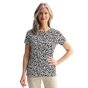 A woman wears a black and white patterned short-sleeve shirt paired with light-colored pants, standing with one hand on her hip and smiling.