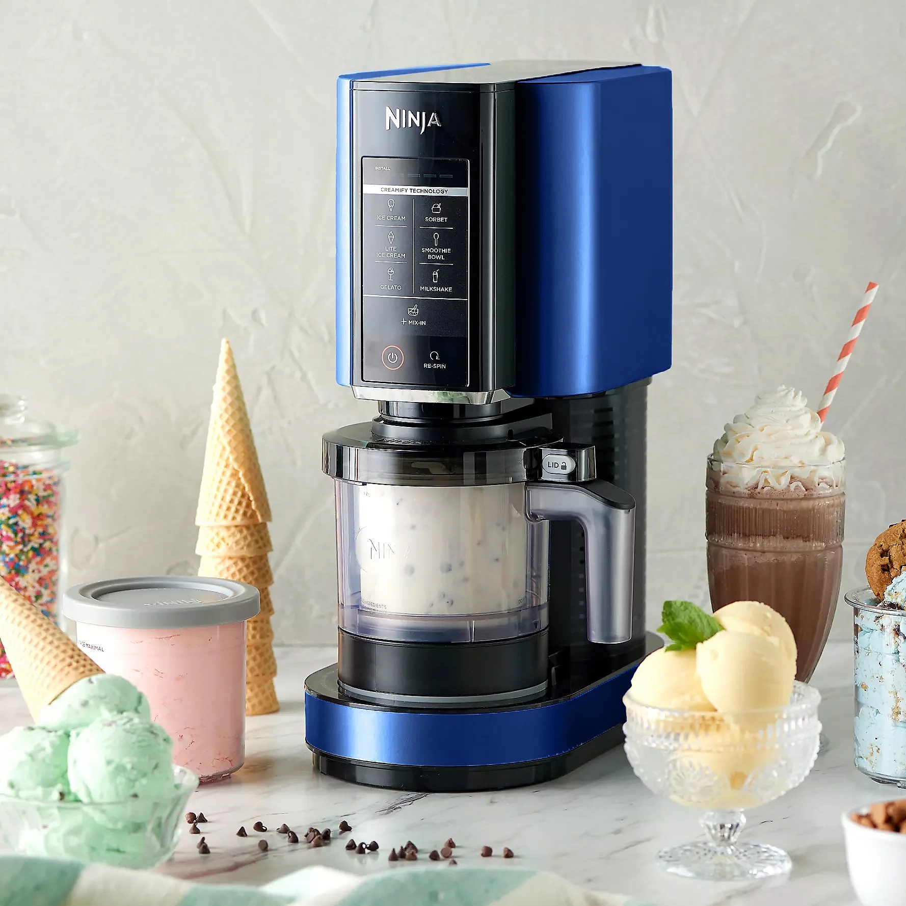 A blue Ninja ice cream maker is displayed on a countertop alongside ice cream, cones, a shake, and sprinkles.