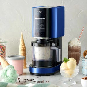 A blue Ninja ice cream maker is displayed on a countertop alongside ice cream, cones, a shake, and sprinkles.