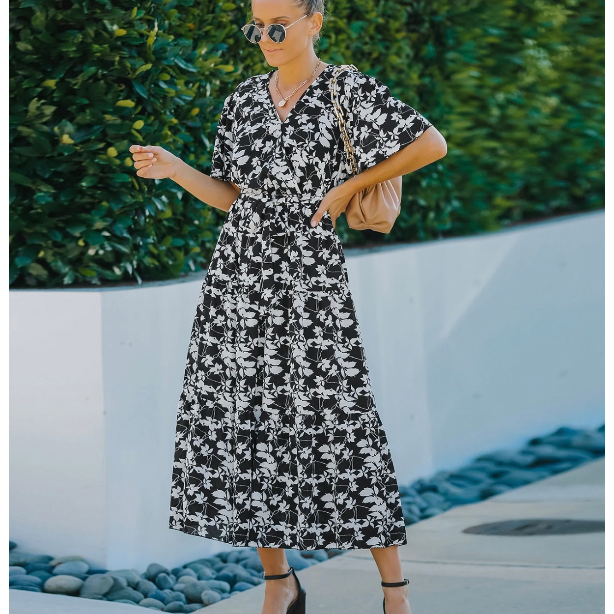 A woman wearing a black and white floral print midi dress with short sleeves and a V-neckline, accessorized with sunglasses, black heels, and a beige shoulder bag.