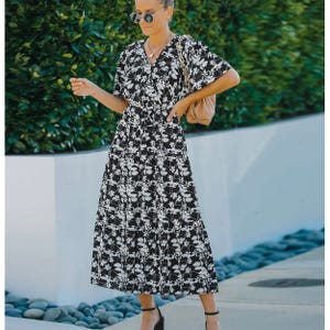 A woman wearing a black and white floral print midi dress with short sleeves and a V-neckline, accessorized with sunglasses, black heels, and a beige shoulder bag.
