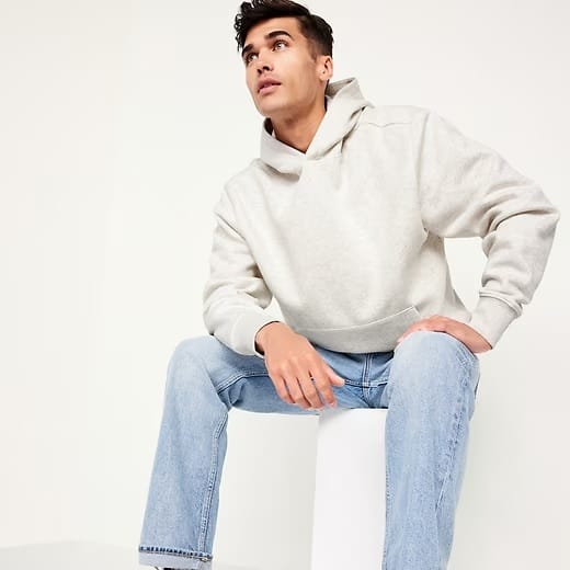 A man wearing a light beige hoodie and light blue jeans sits on a white block.