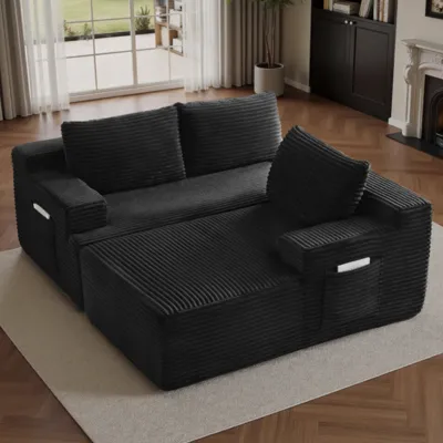 A black, corduroy-textured sofa with a chaise lounge features side pockets for storage, set on a beige rug in a living room with wooden floors and a fireplace.
