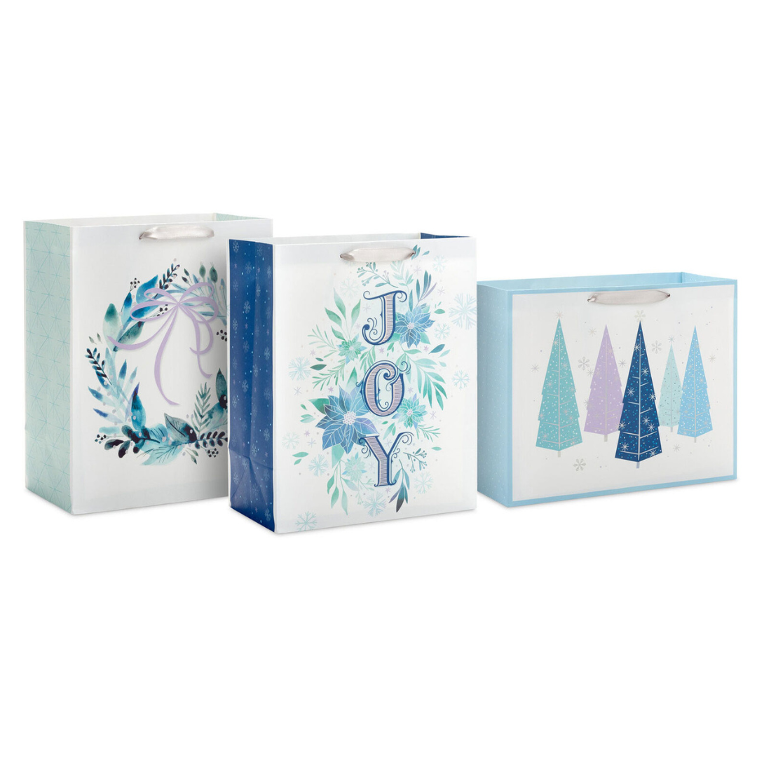 Three festive blue-themed gift bags feature designs with a wreath, the word \“JOY\“ with florals, and abstract Christmas trees, each with ribbon handles.