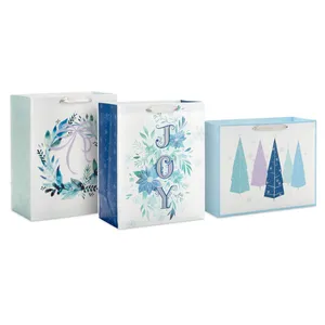 Three festive blue-themed gift bags feature designs with a wreath, the word \“JOY\“ with florals, and abstract Christmas trees, each with ribbon handles.