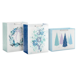 Three festive blue-themed gift bags feature designs with a wreath, the word \“JOY\“ with florals, and abstract Christmas trees, each with ribbon handles.