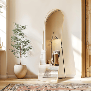 Tall floor mirror with an arched top and thin gold frame leans against a wall next to a large potted plant. The setting includes a patterned rug, light wood flooring, and a glimpse of a cozy room with a lamp and cushioned chair in the reflection.