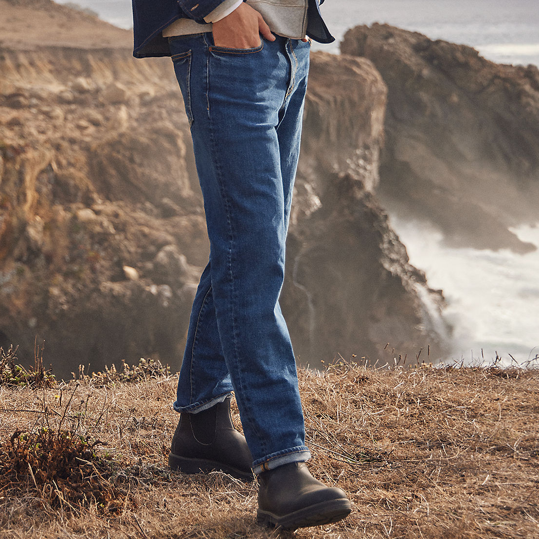Men's straight-leg jeans in a classic blue wash with a five-pocket design, paired with black leather boots. The setting is an outdoor cliffside landscape.