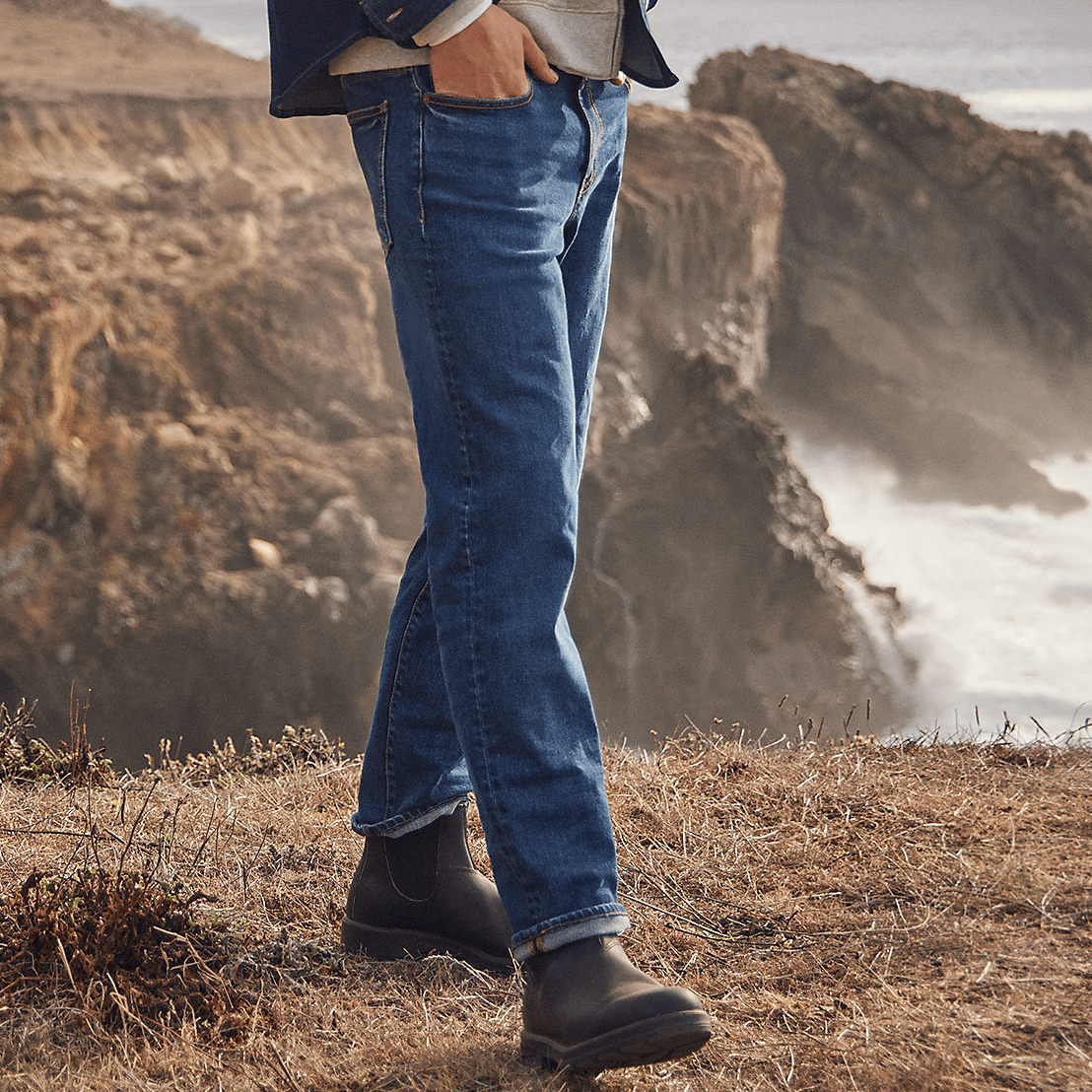 Men's straight-leg jeans in a classic blue wash with a five-pocket design, paired with black leather boots. The setting is an outdoor cliffside landscape.