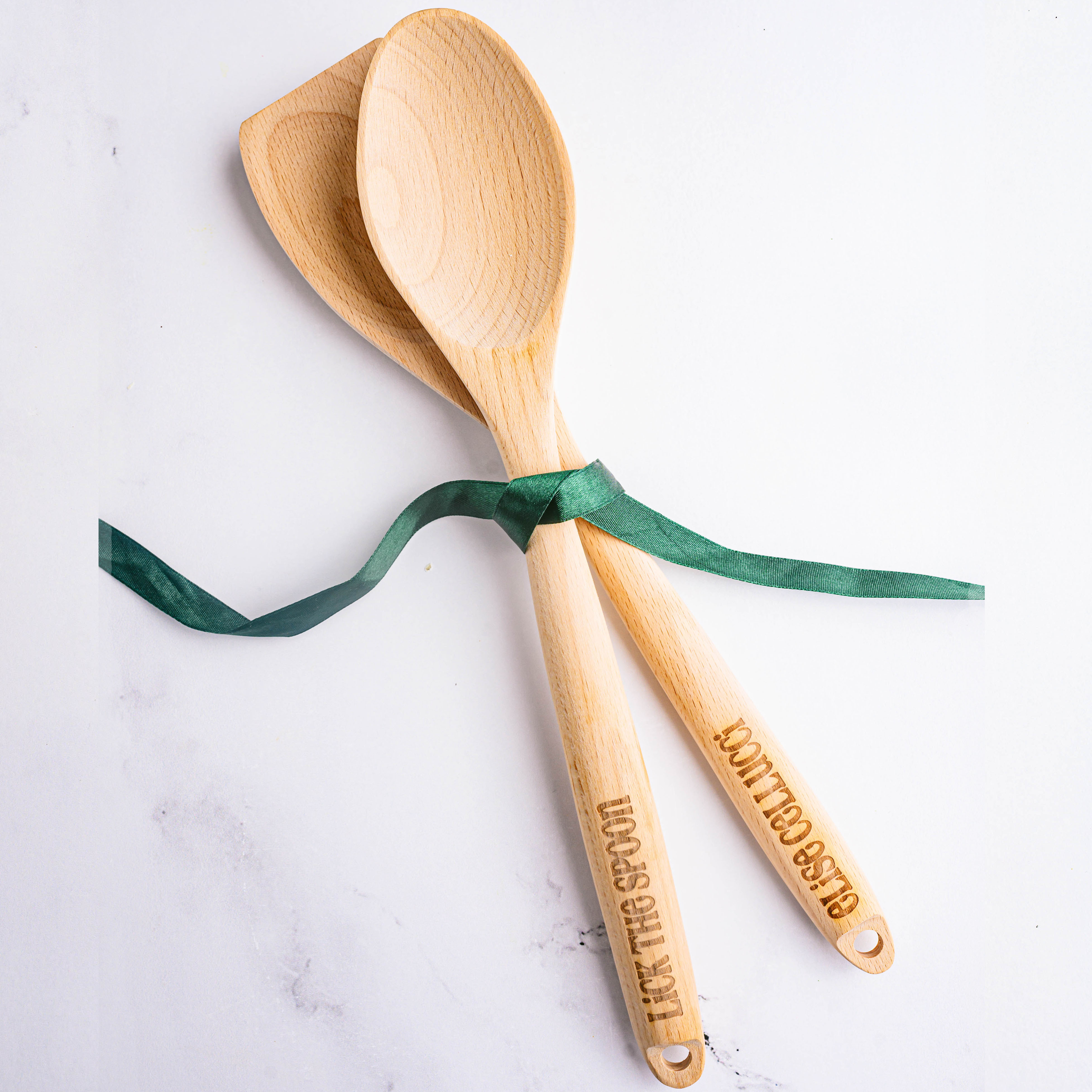 Two wooden kitchen utensils with personalized engraving on the handles are tied together with a green ribbon.