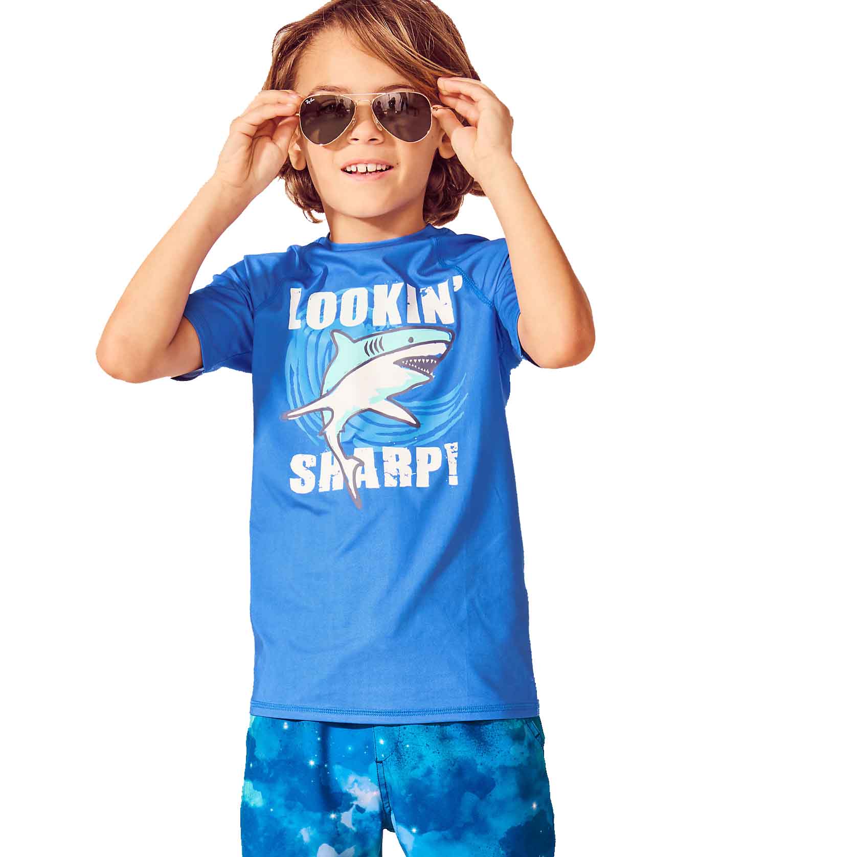 A child wearing sunglasses, a blue shirt with a shark graphic and \“LOOKIN' SHARP!\“ text, paired with blue shorts featuring a water-like pattern.