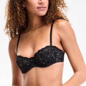 A woman wearing a black lace balconette bra with thin straps and a small bow detail in the center.