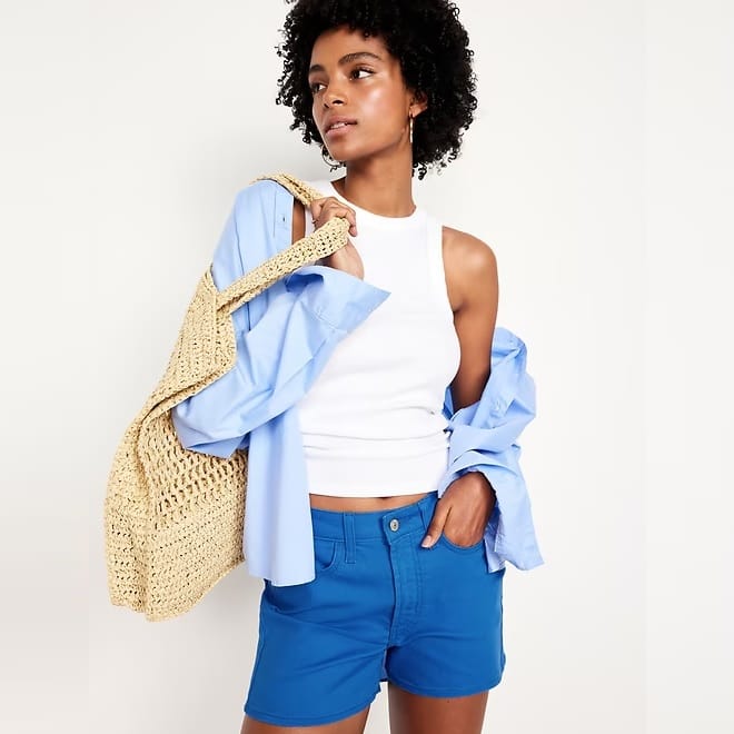 A woman is wearing blue shorts, a white tank top, and holding a light blue shirt and a straw tote bag.
