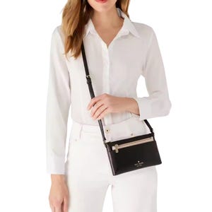 A woman in a white blouse and pants carries a small black Kate Spade New York crossbody bag with a front zip pocket and adjustable strap.